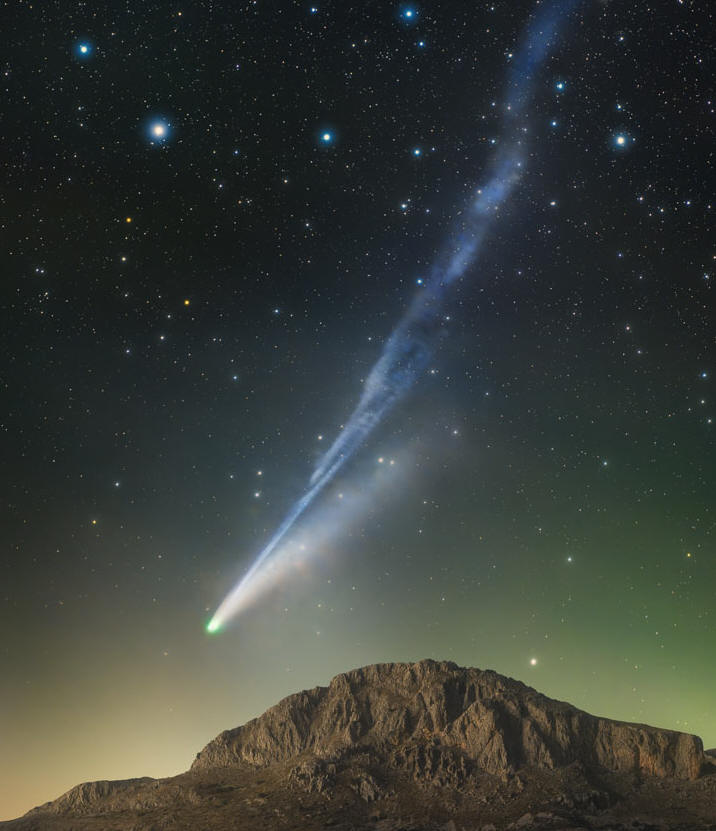 A starfield is shown above a mountain peak. Just
above the mountain and extending up toward the upper 
right is a blue-tinted tail of a comet. The comet's
head is just to the left of the peak.  
Please see the explanation for more detailed information.