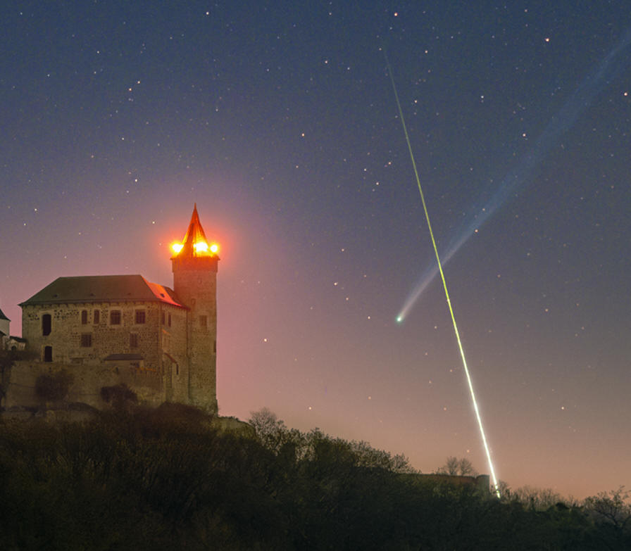 A timelapse image showing a bright streak of light crossing in front of a comet's long tail to form an X-like shape in the night sky. A medieval castle is illuminated in the foreground.